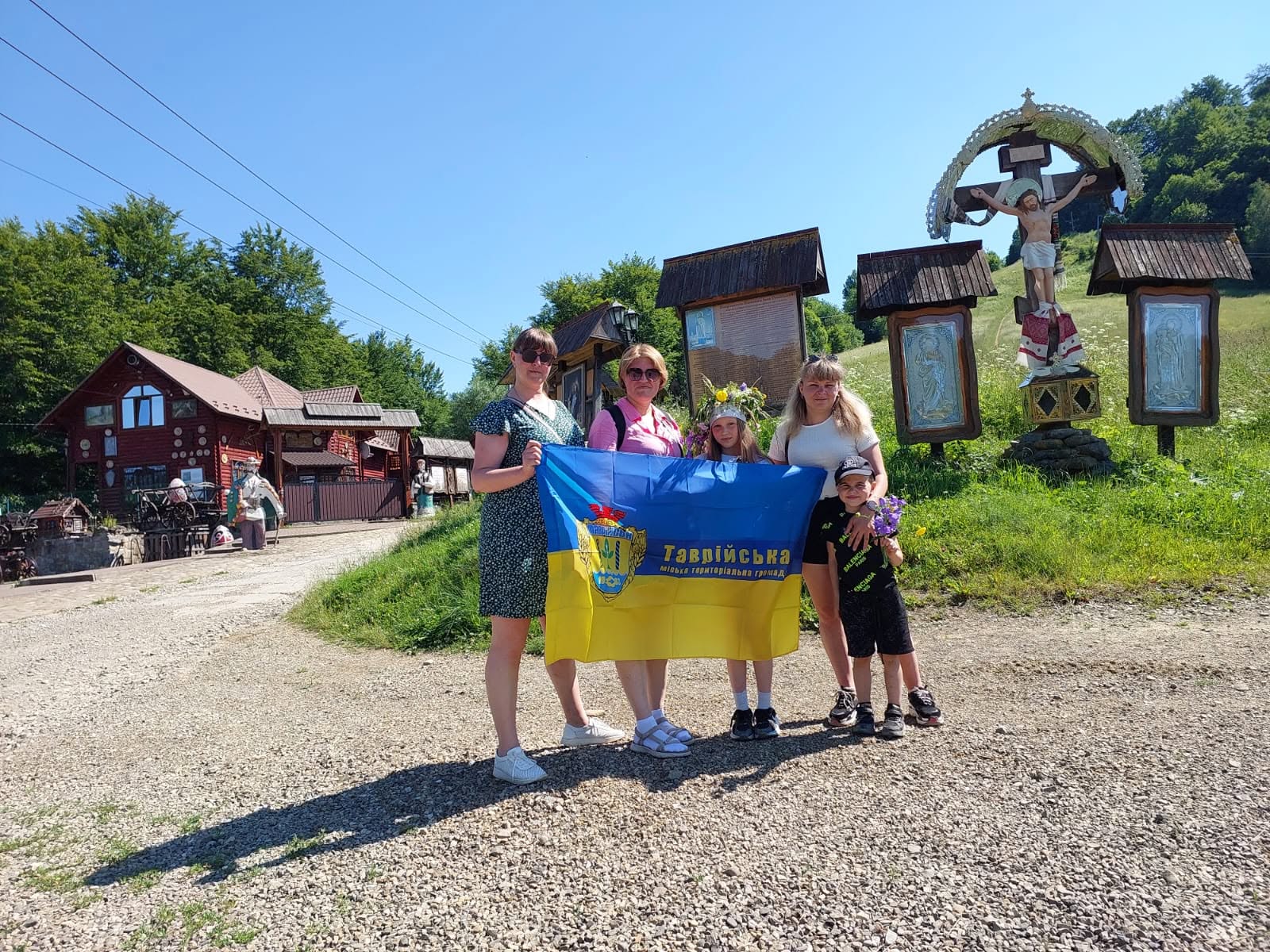 Група з трьох жінок та двох дітей позирує для фото на гравійній дорозі, тримаючи великий синьо-жовтий прапор із написом «Таврійська міська територіальна громада». Вони стоять на тлі дерев'яних релігійних споруд із розп'яттям та іконами, а ліворуч на задньому плані видно коричневі дерев'яні будинки та зелені дерева.