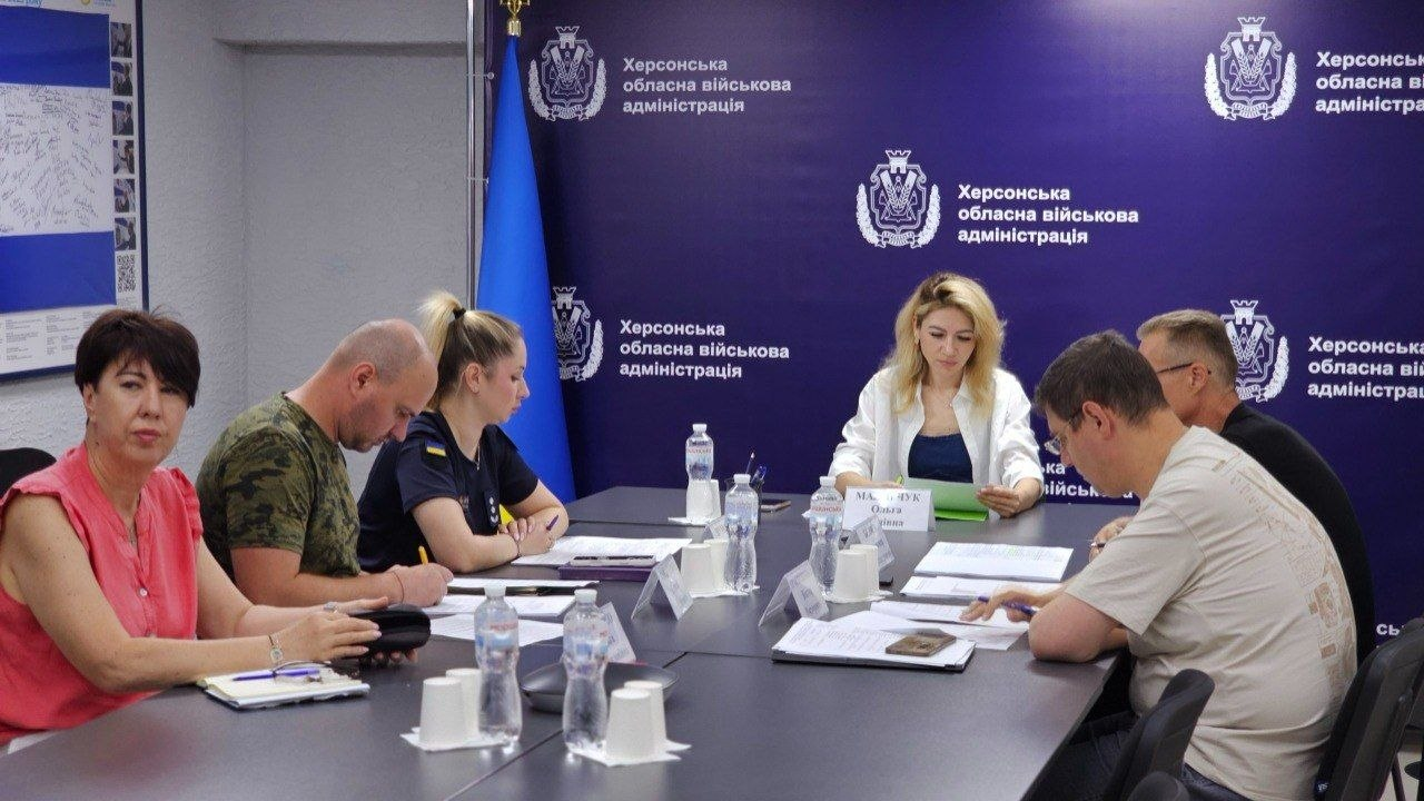 Учасники офіційного засідання — чоловіки та жінки у цивільному одязі, камуфляжних футболках та формі — сидять за великим Т-подібним столом і зосереджено працюють із документами. На столі розкладені папки, блокноти, ручки. Зустріч проходить у робочому кабінеті на тлі синього банера з офіційною символікою, ліворуч від якого стоїть Державний Прапор України.