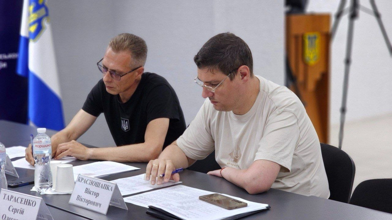 Двоє чоловіків в окулярах та футболках сидять за столом під час офіційного заходу та зосереджено вивчають документи. На столі перед ними лежить мобільний телефон та розміщені настільні таблички з іменами. На задньому плані видно Державний Прапор України та дерев'яну трибуну з гербом.