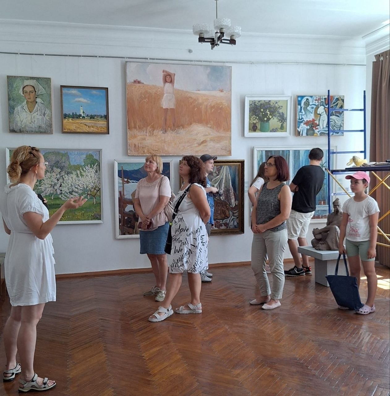 Група відвідувачів у літньому одязі слухає екскурсовода у залі художнього музею з паркетною підлогою. На білих стінах розвішано різноманітні картини, серед яких пейзажі та портрети, а праворуч на постаменті стоїть скульптура.