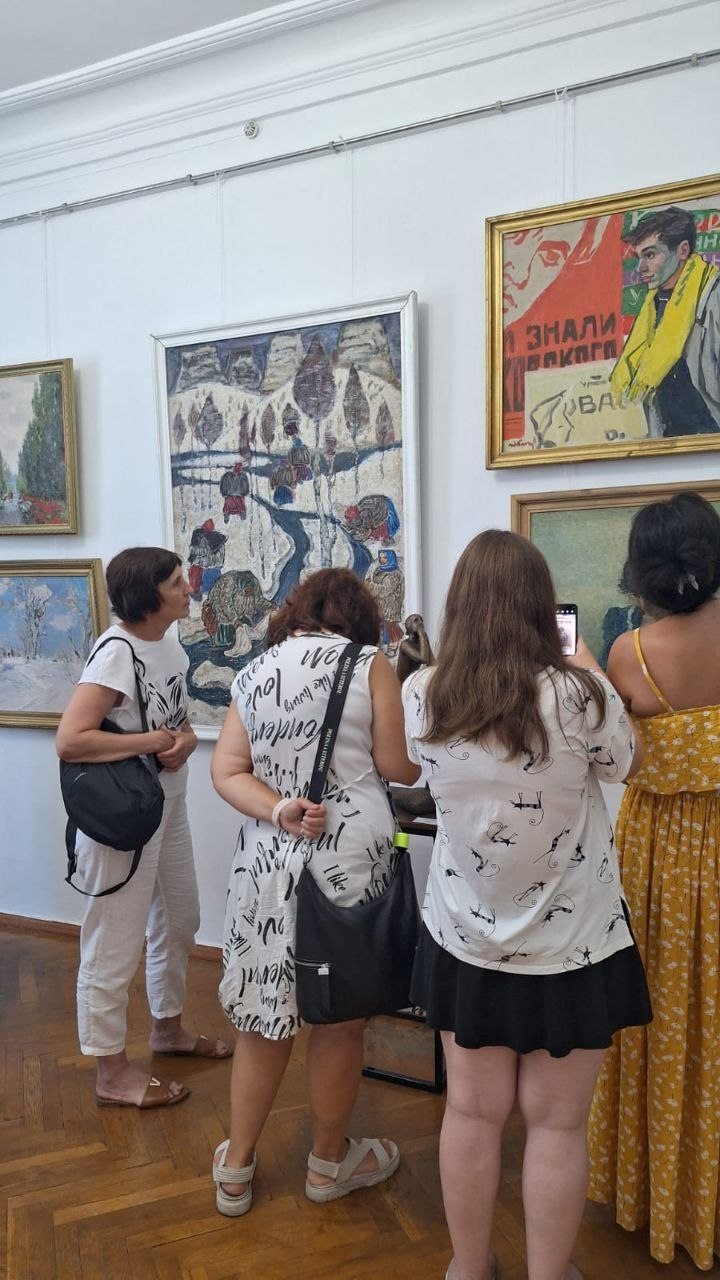 Чотири жінки стоять біля стіни в музейному залі та розглядають розвішані картини в рамах. Одна з відвідувачок фотографує експозицію на свій мобільний телефон.