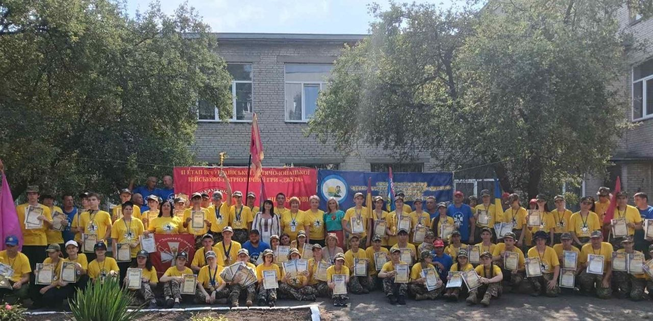 Велика група молоді та дорослих у жовтих футболках із дипломами в руках позує для спільного фото на подвір'ї будівлі на тлі банерів патріотичної гри «Джура».