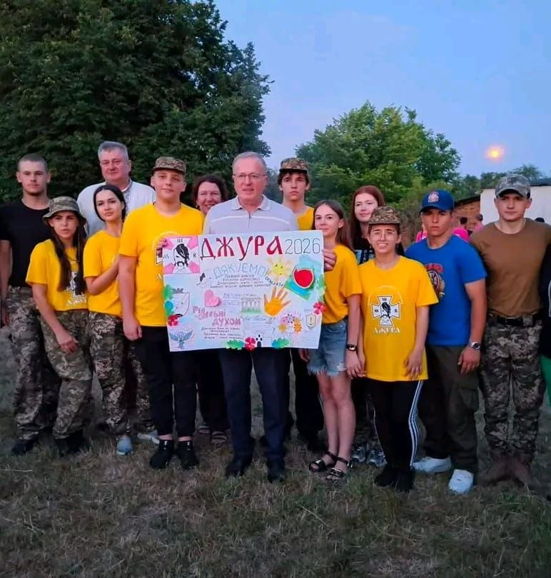 Група молоді у жовтих футболках та камуфляжних штанах разом із дорослими стоїть на вулиці у вечірній час і тримає великий саморобний плакат із написами «Джура 2026» та «Дякуємо», прикрашений малюнками й дитячими побажаннями.