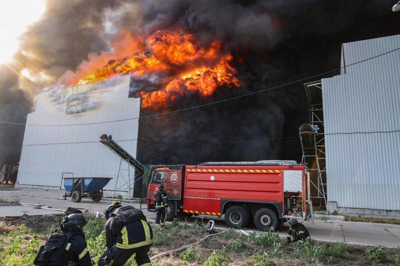 Масштабна пожежа на промисловому підприємстві, де вогонь повністю охопив дах великої білої будівлі з металопрофілю. У небо здіймається величезна стіна інтенсивного помаранчевого полум'я та густого чорного диму. На передньому плані стоїть червоний пожежний автомобіль, а рятувальники в захисному спорядженні готуються до гасіння вогню.