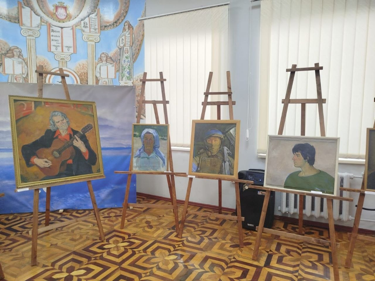 Виставка живопису в приміщенні з паркетною підлогою, де на дерев'яних мольбертах представлено п'ять різних картин. Серед полотен — портрет чоловіка з гітарою, портрет жінки у хустці, портрет військовослужбовця у спорядженні та портрет хлопця у профіль. На задньому плані видно вікна з жалюзі та стіну з великим художнім розписом.
