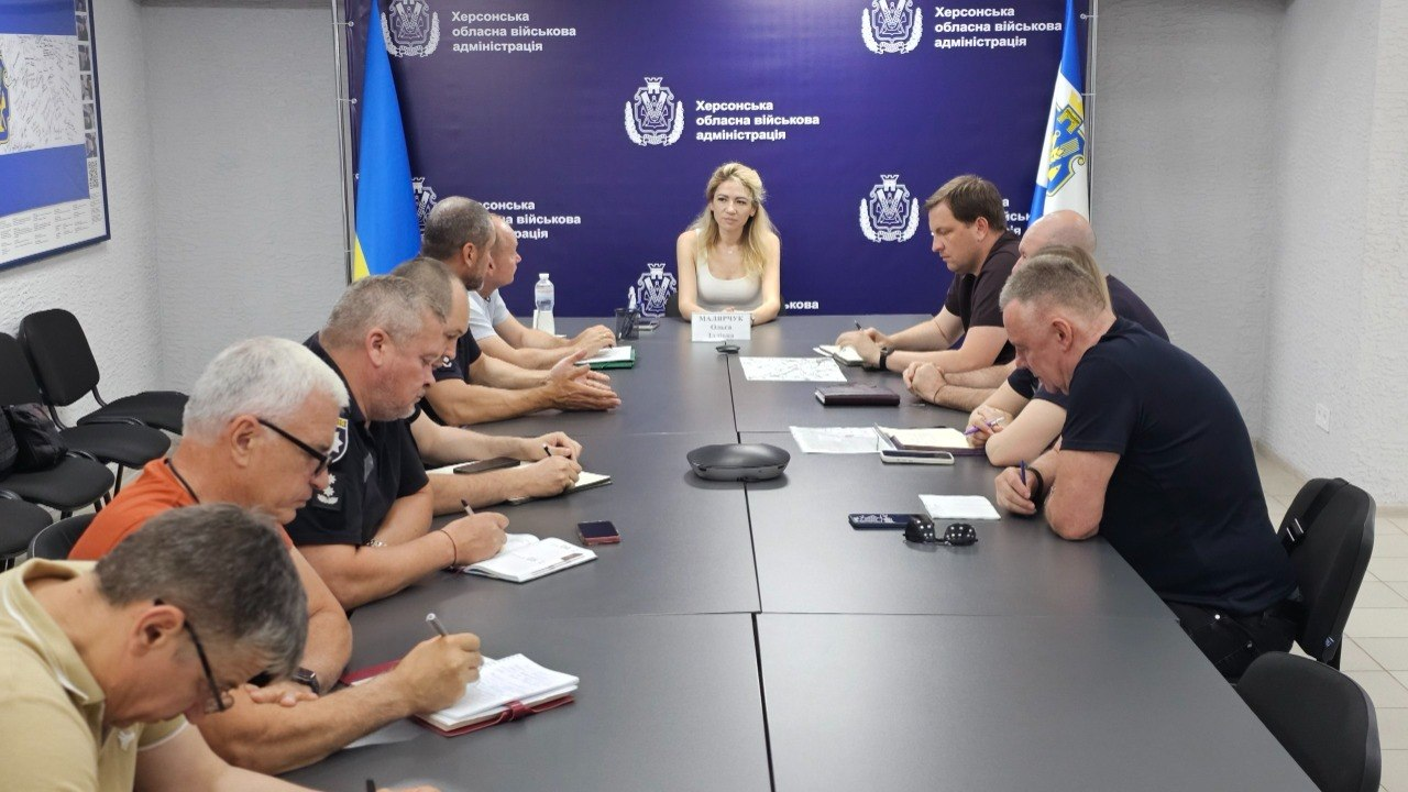 Група людей у цивільному та службовому одязі проводить робочу зустріч за довгим сірим Т-подібним столом. Учасники роблять нотатки, вивчають документи або слухають жінку, яка сидить на чолі столу. На задньому плані видно синій банер із гербами та два державні прапори України.