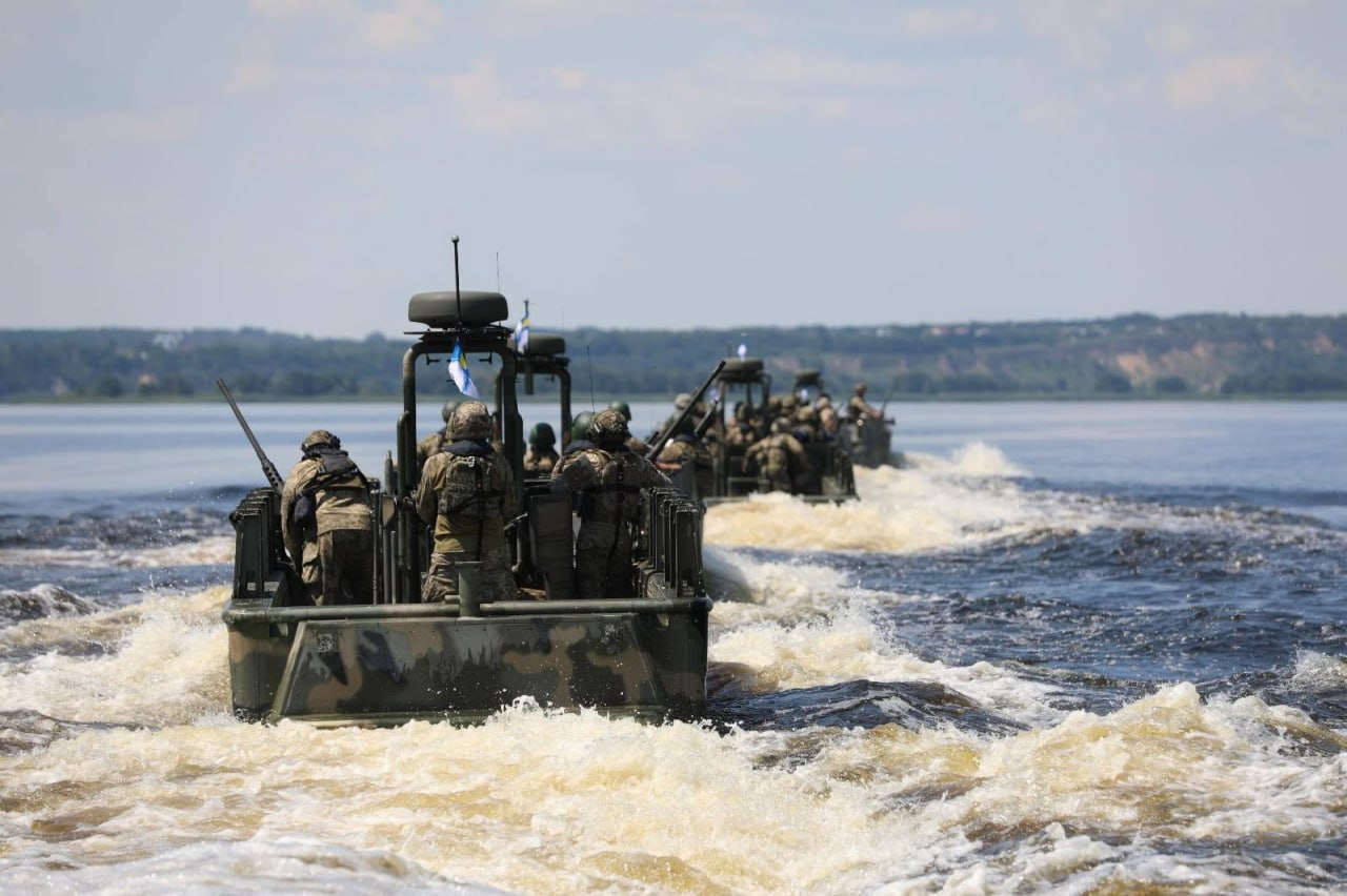 Колона військових швидкісних катерів рухається по водній поверхні, залишаючи за собою білі пінисті хвилі. На борту човнів перебувають військовослужбовці у повній екіпіровці, камуфляжній формі та шоломах. На задньому плані видно широку річку або водосховище з розмитим лінією берега під хмарним небом.