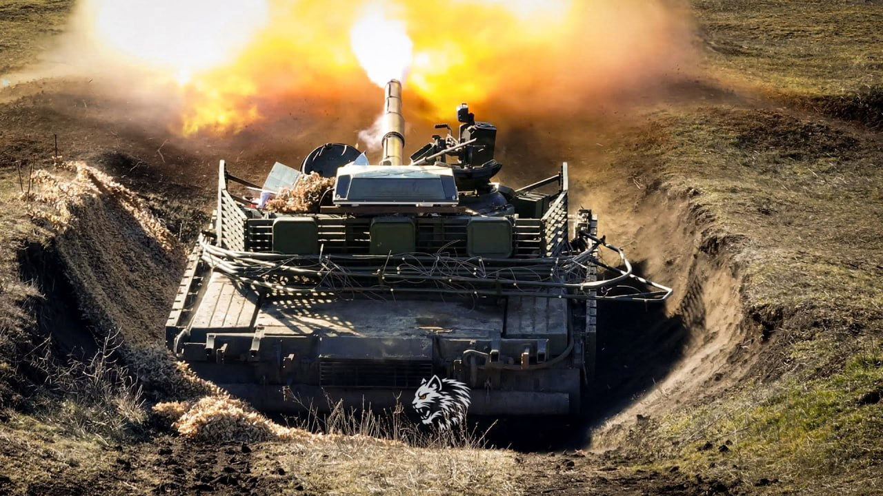 Військовий танк здійснює постріл з окопу або укриття, у момент якого з дула виривається великий спалах вогню та диму. Техніка заглиблена в землю, видно деталі її корпусу та захисних решіток ззаду. На передньому плані по центру розміщено невеликий білий логотип із зображенням вовка.