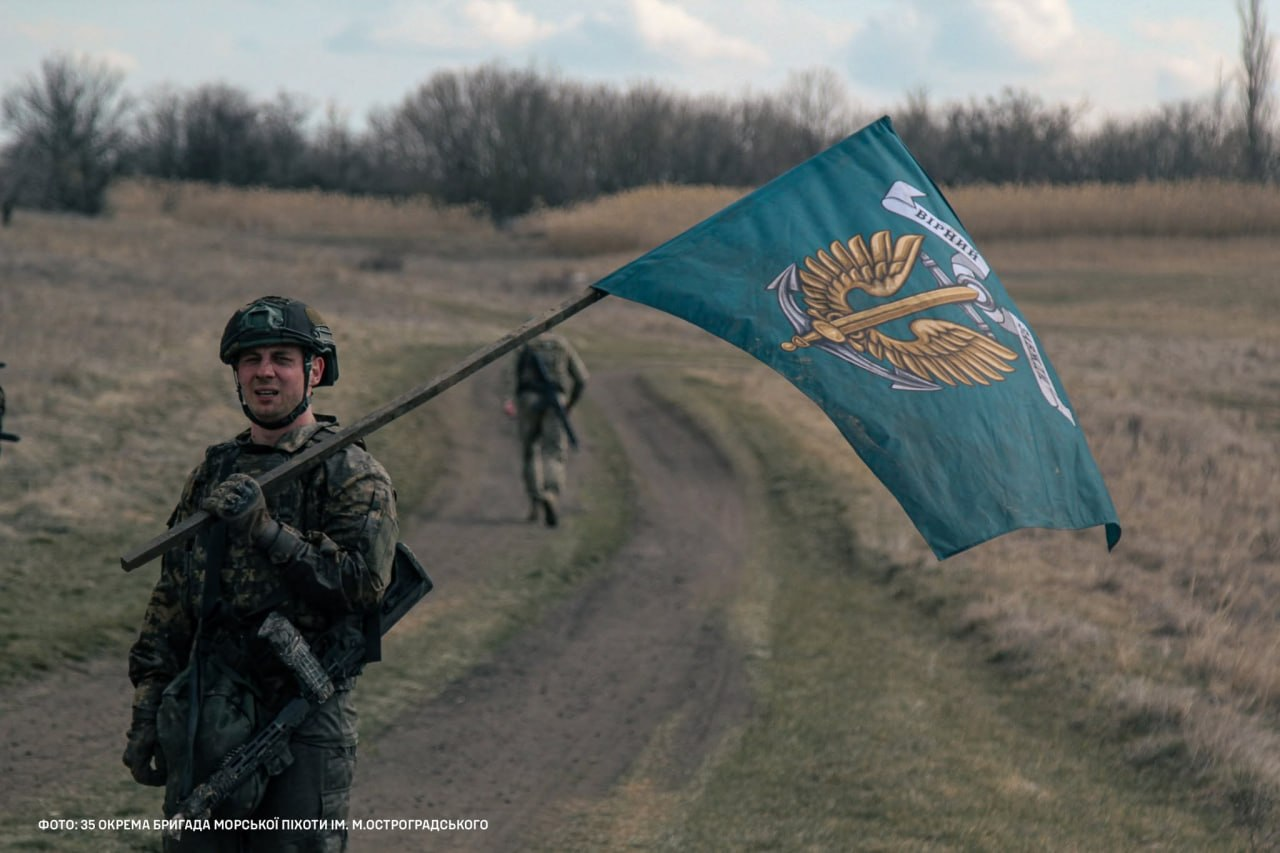 Військовослужбовець у камуфляжній формі, бронежилеті та шоломі стоїть на роздоріжжі польової дороги, тримаючи на плечі великий бірюзовий прапор Морської піхоти України. На задньому плані видно іншого військового, який іде дорогою вглиб поля, та сухі чагарники з деревами під хмарним небом.