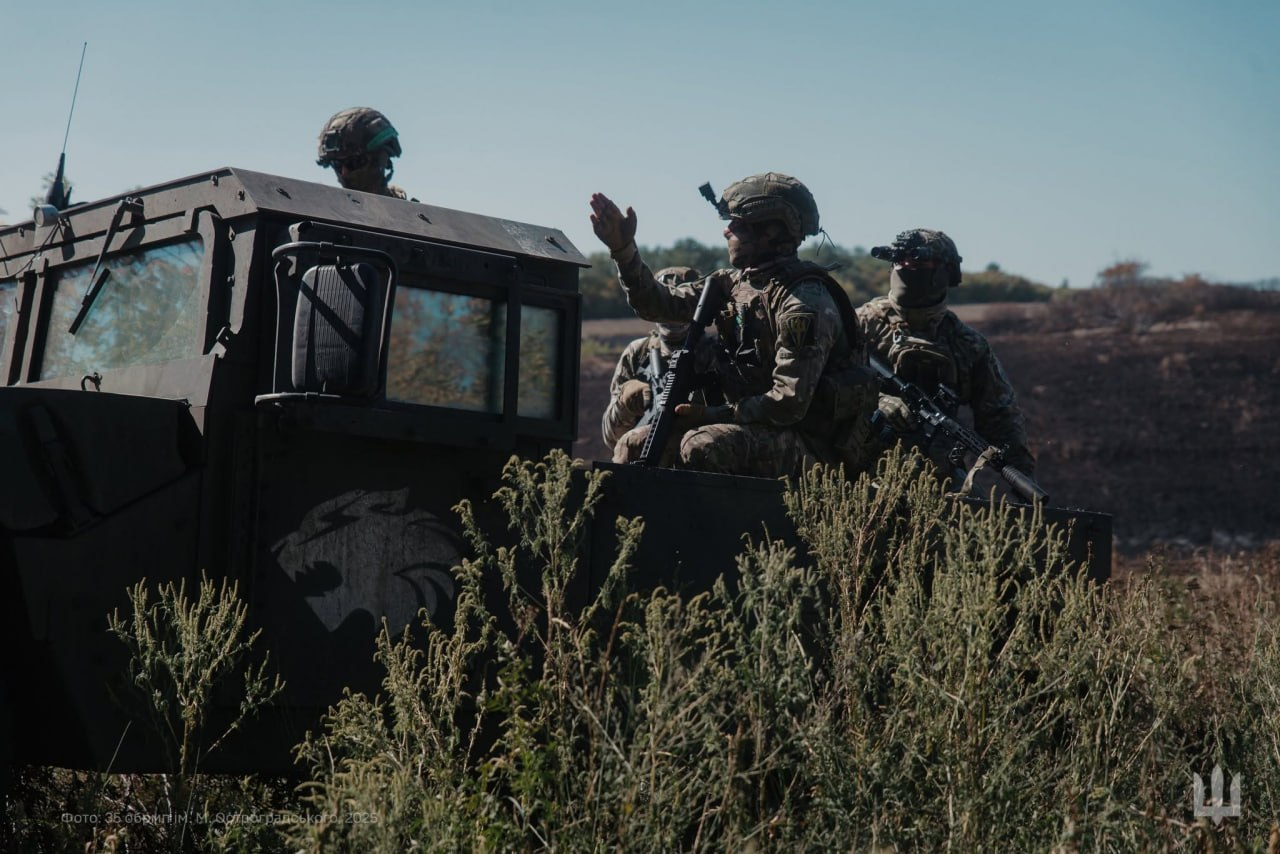 Військовослужбовці у повній екіпіровці, шоломах та зі зброєю перебувають у кузові та кабіні темного бронеавтомобіля серед високої сухої трави. Один із бійців підняв руку, вказуючи напрямок або подаючи сигнал іншим. На борту машини видно сіре зображення голови хижака, а в кутку кадру — невеликий білий тризуб.