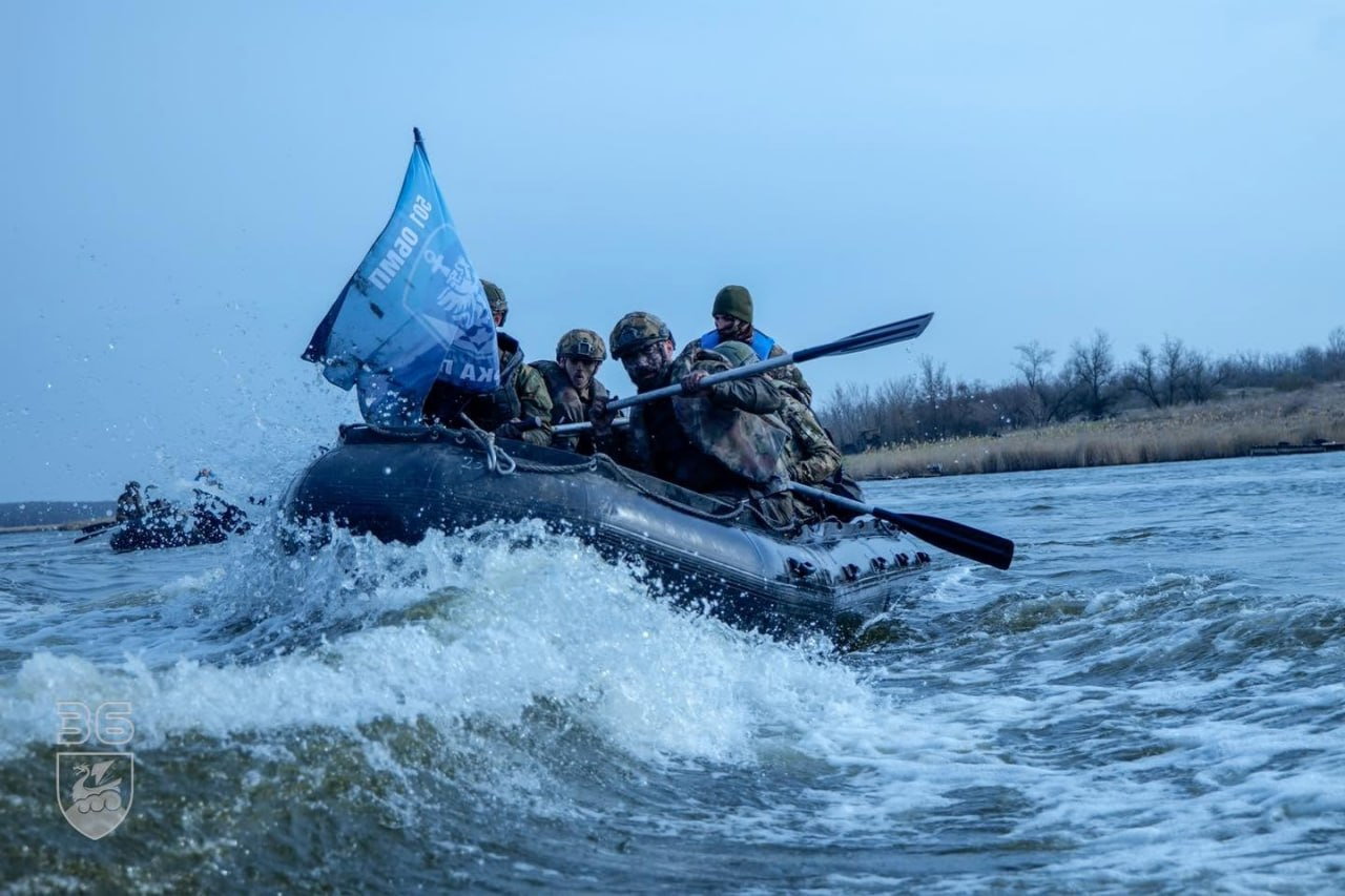 Група військовослужбовців у захисному спорядженні та шоломах пличе на надувному моторному човні по річці, долаючи сильні хвилі та бризки. На носі човна встановлено блакитний прапор із символікою Морської піхоти України. На задньому плані видно інший човен та пустельний берег під похмурим небом.
