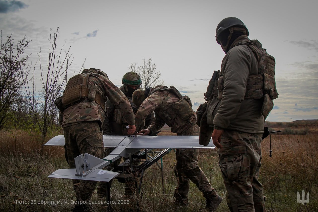 Четверо військовослужбовців у захисному спорядженні та камуфляжі готують до запуску безпілотний літальний апарат літакового типу на стартовій катапульті. Один боєць стоїть праворуч і спостерігає, а троє інших нахилилися над безпілотником серед сухої трави. У лівому нижньому кутку є дрібний напис із копірайтом, а праворуч — білий символ тризуба.