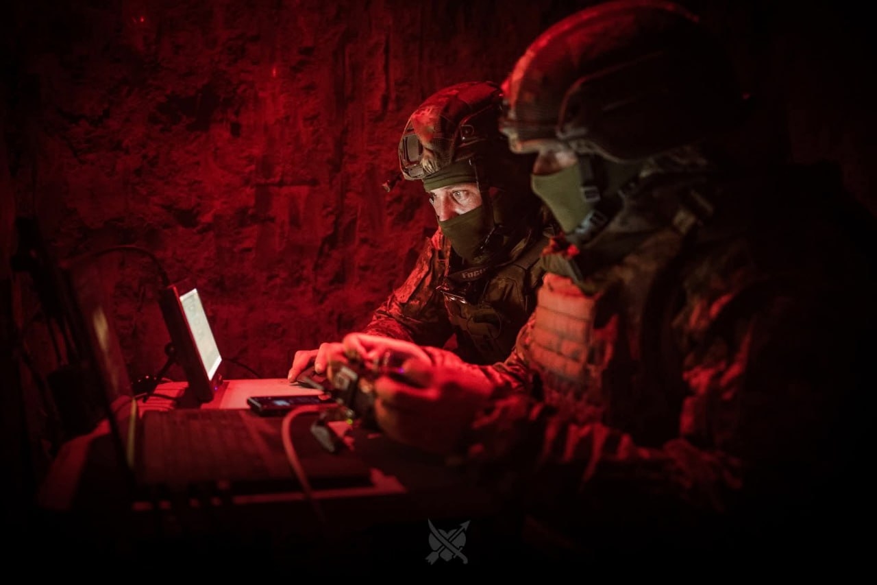 Двоє військовослужбовців у шоломах та захисних масках працюють у темному бліндажі або укритті під червоним тактичним освітленням. Вони сидять перед екранами ноутбука та монітора, керуючи обладнанням за допомогою пульта або джойстика. У нижній частині кадру по центру ледь помітний сірий логотип із перехрещеними мечами.