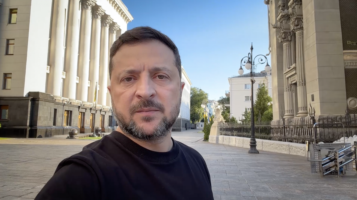 Чоловік у чорному світшоті знімає селфі-відео на фоні урядової будівлі з колонами та Будинку з химерами в Києві. На вулиці сонячно та безлюдно.