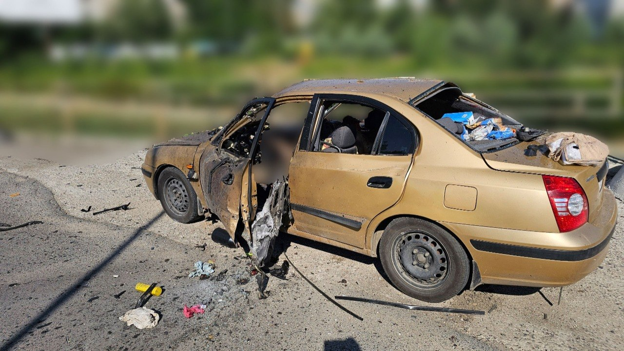 Золотистий легковий автомобіль із серйозними пошкодженнями після вибуху або обстрілу стоїть на асфальтованій дорозі. Передні двері з боку водія розірвані та обгорілі, шибки вибиті, а всередині салону помітні сліди руйнувань та речі. Навколо машини на дорозі розкидані уламки та сміття.