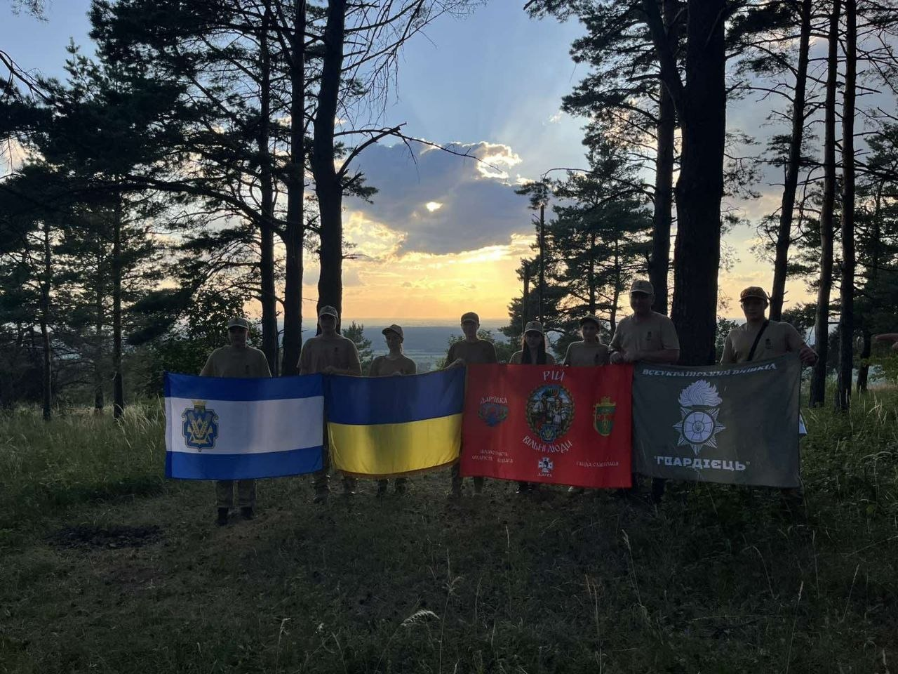 Група людей у формі стоїть на узліссі на тлі вечірнього сонця, що заходить, та тримає чотири різні прапори. Серед них розгорнуто синьо-жовтий прапор України, прапор Херсонської області, червоний прапор із символікою та написами, а також сірий прапор із емблемою та написом «Гвардієць».