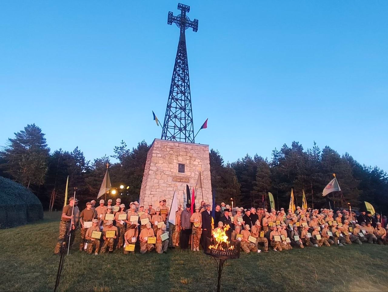 Велика група молоді у військово-патріотичній формі вишикувалася для спільного фото на галявині біля високого кам'яного монумента з металевою вежею та хрестом. Багато учасників тримають у руках грамоти чи дипломи, по центру стоять прапори, а на передньому плані горить вогонь у металевій чаші. На задньому плані височіє густий хвойний ліс під чистим вечірнім небом.