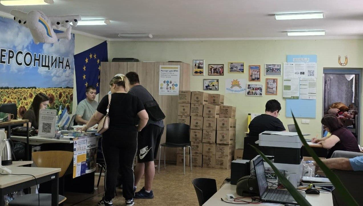 Люди перебувають у приміщенні гуманітарного центру. Кілька відвідувачів стоять біля столу реєстрації, а працівники заповнюють документи та працюють за комп'ютерами. На стіні висить великий банер із соняшниковим полем та прапор Європейського Союзу, а на задньому плані складено багато картонних коробок із допомогою.