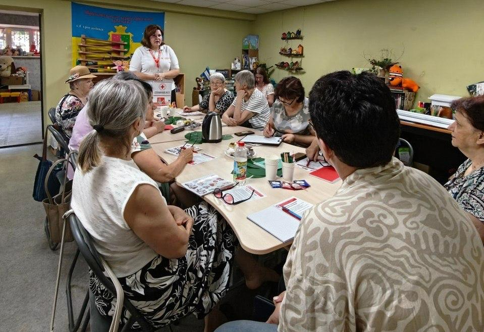 Група людей сидить за великим Т-подібним столом під час зустрічі чи тренінгу в Громадському центру Українського Червоного Хреста. Наприкінці столу стоїть жінка-модератор у білій футболці-поло та розмовляє з присутніми. На столі розкладено блокноти, інформаційні матеріали, ручки, стаканчики та термос, а на стінах кімнати видно полиці з іграшками та банер громади.