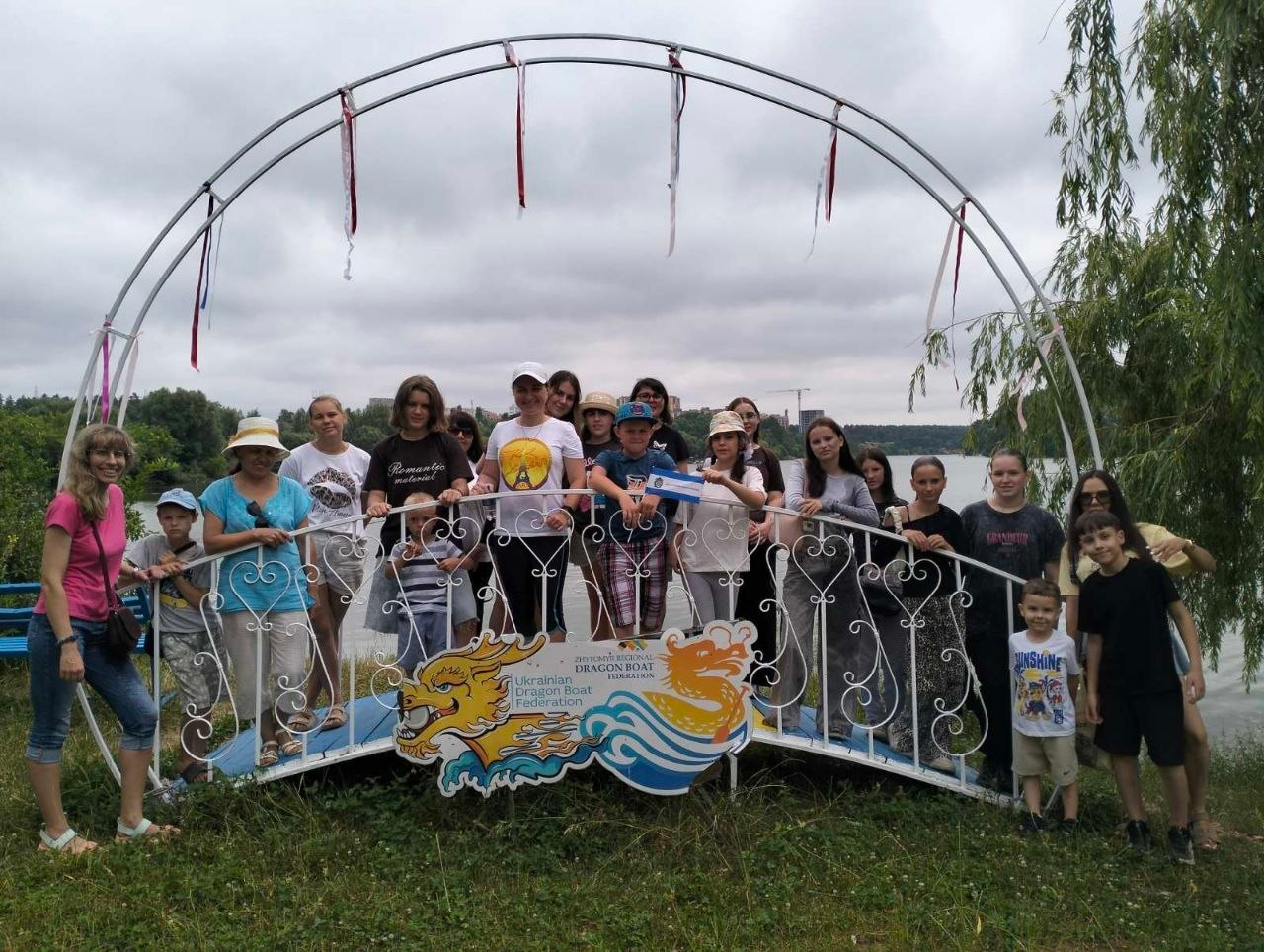 Велика група дітей та дорослих позує для спільного фото на декоративному металевому містку з аркою на березі річки. На передній частині містка закріплена емблема з драконом та написом «Ukrainian Dragon Boat Federation». Учасники посміхаються на тлі широкої водойми, лісу та хмарного неба, а з правого боку фото видно гілки верби.