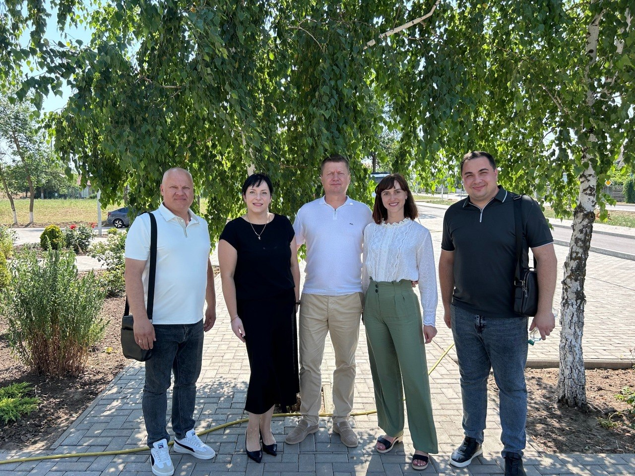Група з п'яти людей — троє чоловіків і дві жінки — стоїть у ряд на вибрукуваній доріжці та посміхається, позуючи для фото під розлогим зеленим деревом у сонячний день. Вони вдягнені в літній повсякденний одяг світлих і темних тонів, а на задньому плані видно міську вулицю з дорогою та іншими зеленими насадженнями.