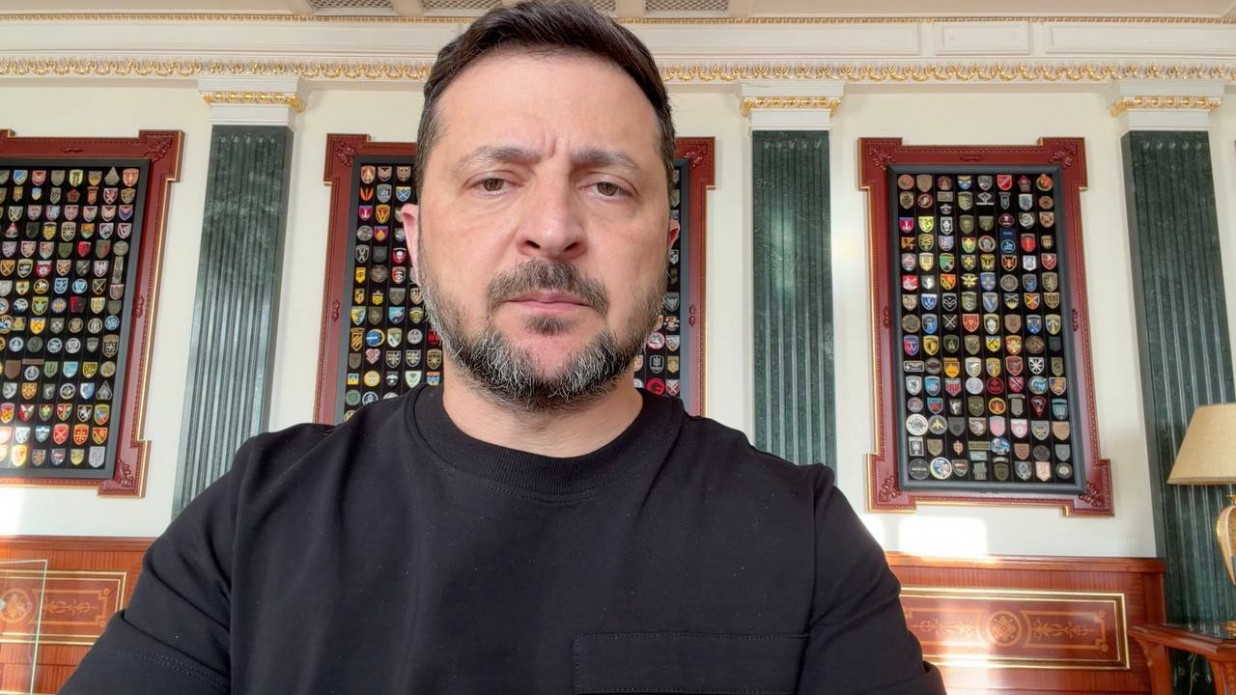Чоловік у чорному світшоті зафіксований крупним планом під час звернення на тлі офіційного кабінету. На стіні позаду нього висять великі стенди, повністю заповнені різноманітними військовими шевронами та емблемами підрозділів.