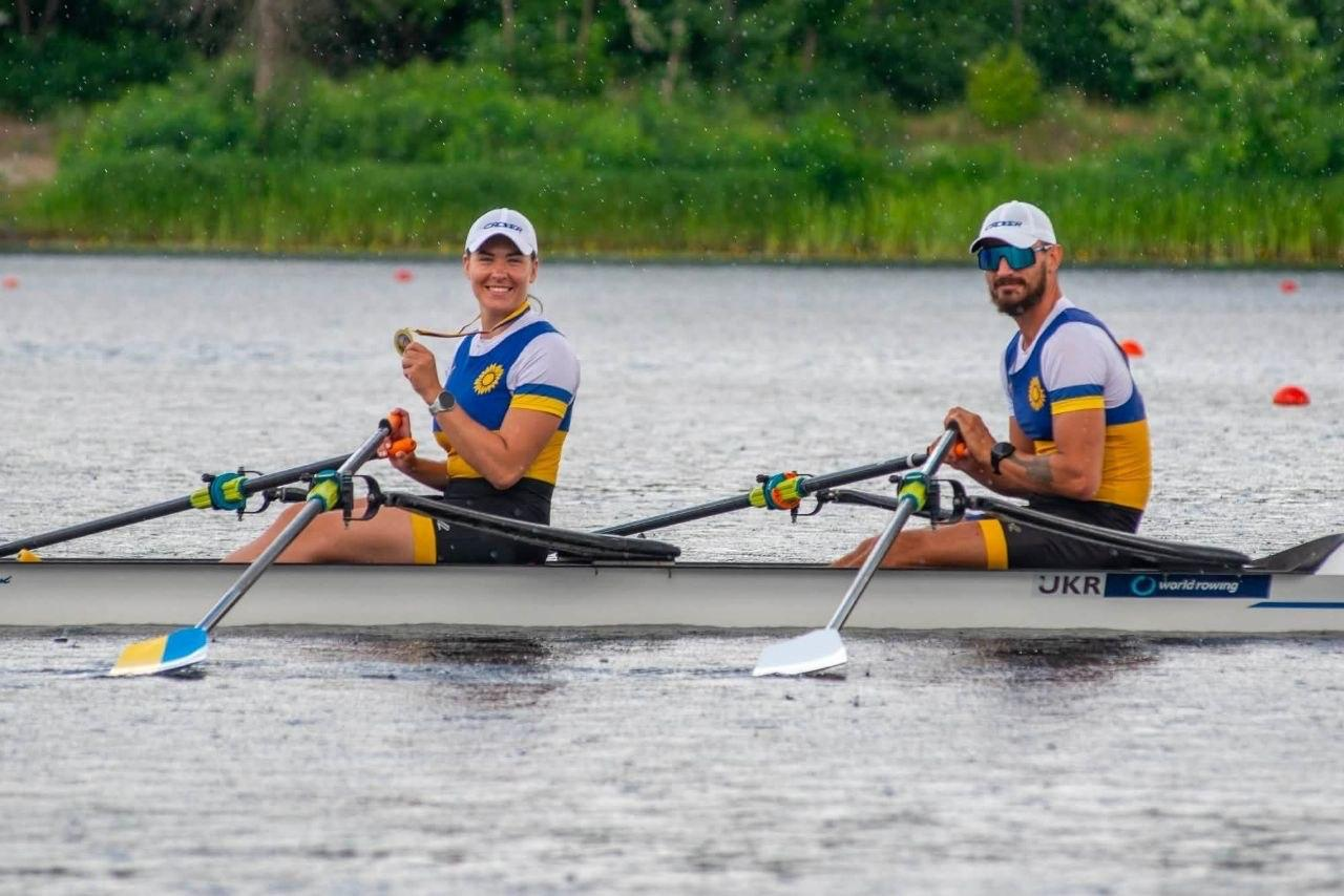 Чоловік та жінка в українській спортивній формі сидять у двомісному човні для академічного веслування на воді, при цьому жінка посміхається та тримає в руці медаль.