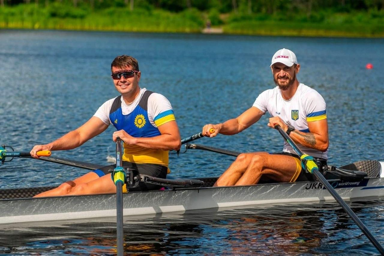 Двоє чоловіків в українській спортивній формі посміхаються, сидячи у двомісному човні для академічного веслування на відкритій водоймі.