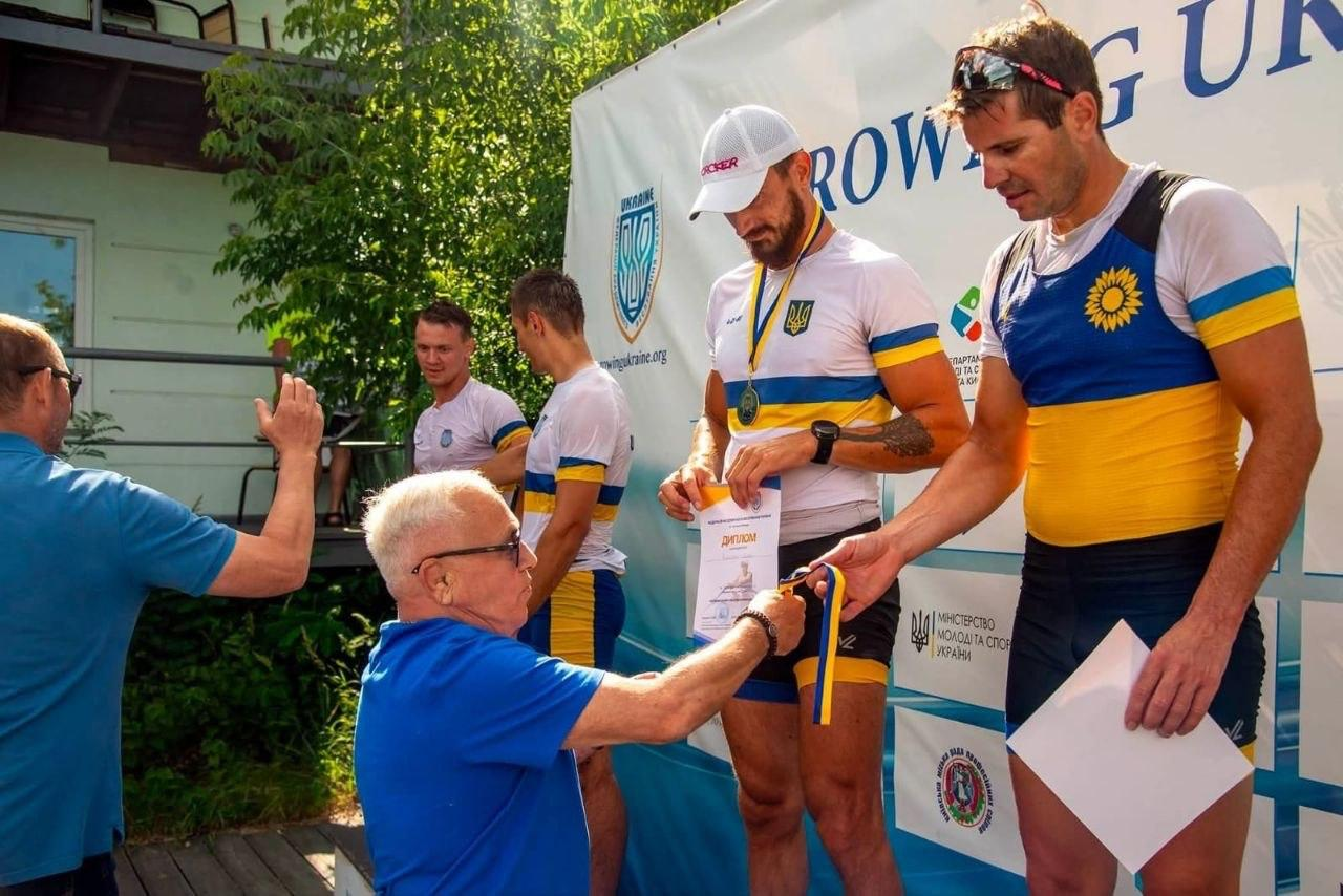 Літній чоловік у синій футболці одягає медаль на шию спортсмену в синьо-жовтій формі під час офіційного нагородження.