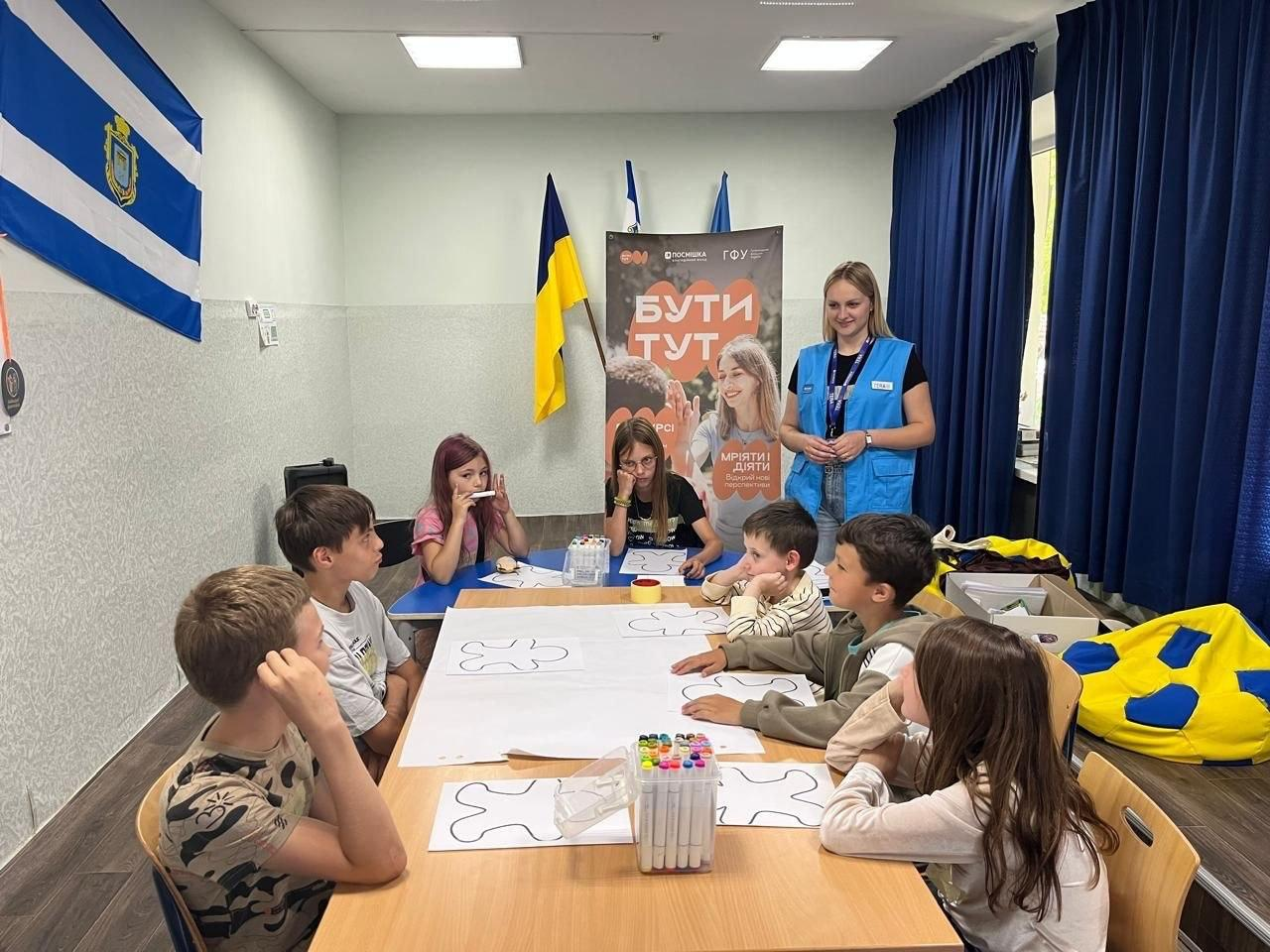 Група дітей сидить за великим столом і малює, а поруч стоїть жінка-волонтер у синьому жилеті на тлі прапорів та банера з написом «Бути тут».