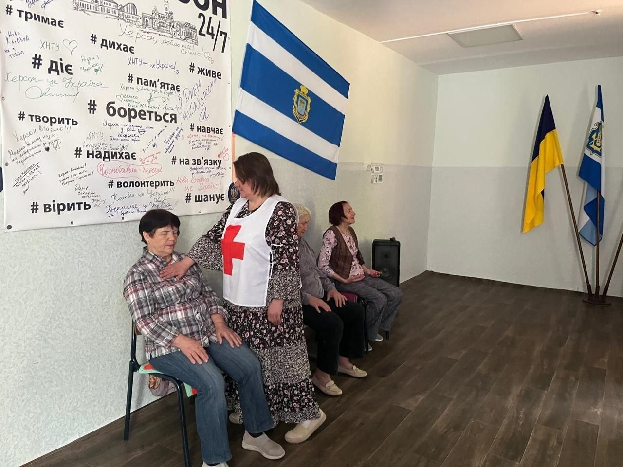 Група людей сидить на стільцях під час заходу в приміщенні, де одна з жінок одягнена у медичний жилет із червоним хрестом, на тлі стіни з патріотичними написами та прапорами.