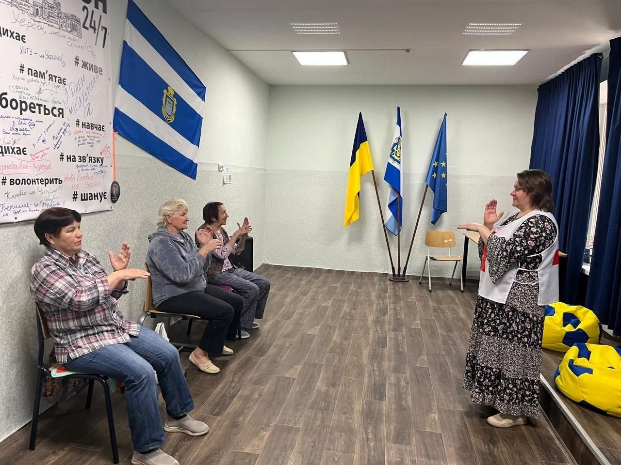 Жінка у жилеті Червоного Хреста стоїть перед групою людей, які сидять на стільцях та аплодують, у кімнаті з прапорами та патріотичними написами на стінах.
