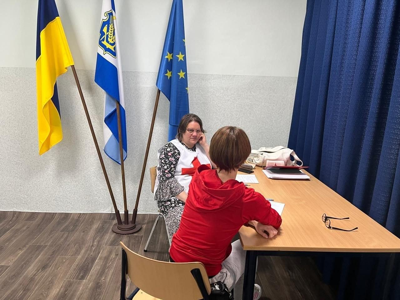Жінка у жилеті Червоного Хреста сидить за робочим столом і спілкується з відвідувачкою у червоній кофті на тлі прапорів України, міста Херсон та Європейського Союзу.
