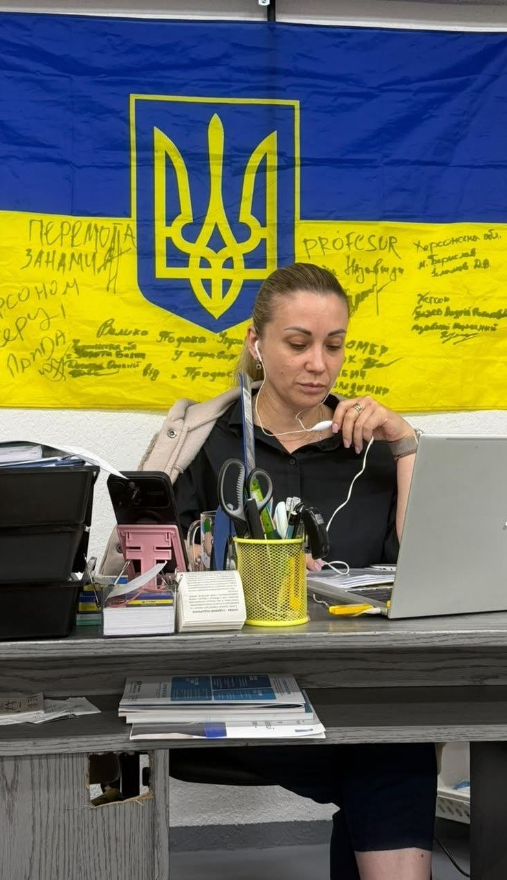 Жінка в навушниках працює за ноутбуком біля робочого столу на тлі великого прапора України з написами від руки та гербом по центру.
