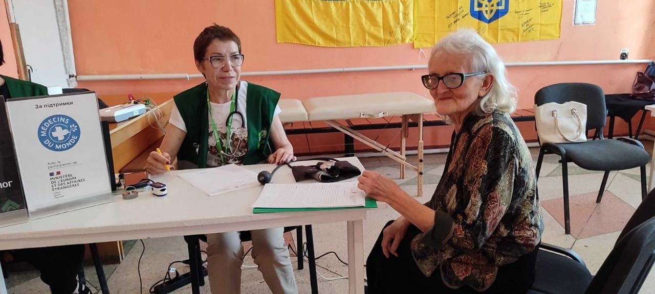 Жінка-медик у зеленому жилеті «Médecins du Monde» із тонометром на столі консультує літню жінку в окулярах у приміщенні з українським прапором на стіні.