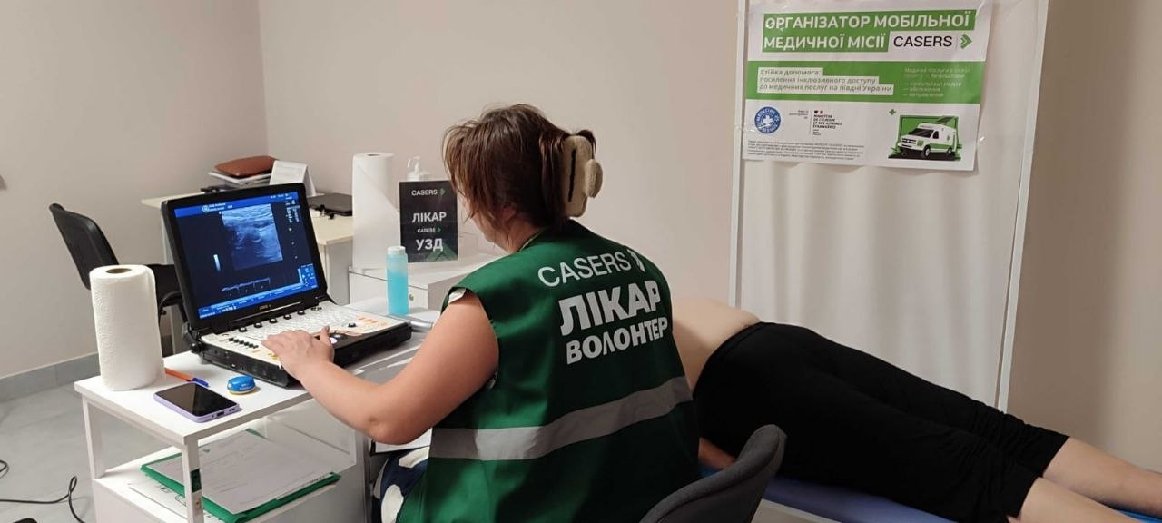 Жінка-медик у зеленому жилеті з написом «CASERS ЛІКАР ВОЛОНТЕР» проводить ультразвукове дослідження (УЗД) пацієнту, який лежить на кушетці в кабінеті.