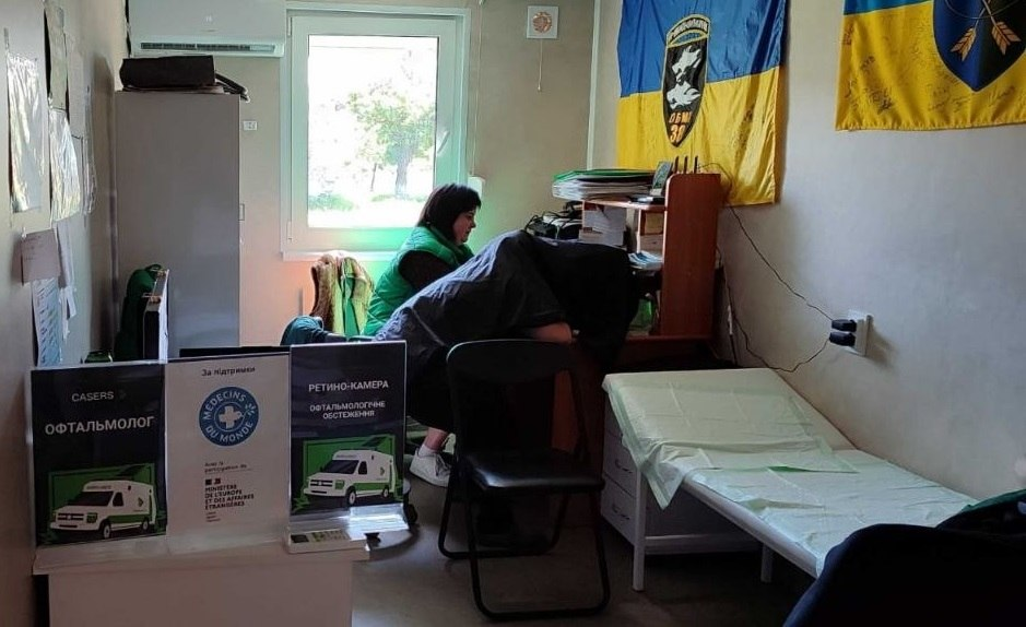 Лікар-офтальмолог проводить обстеження пацієнта за допомогою ретино-камери в медичному кабінеті з українськими прапорами на стіні.