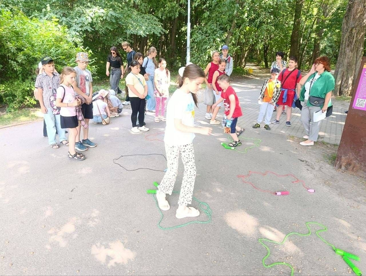 Група дітей разом із дорослими бере участь у рухливих іграх з наочними контурами на асфальтованій доріжці серед дерев у парку.
