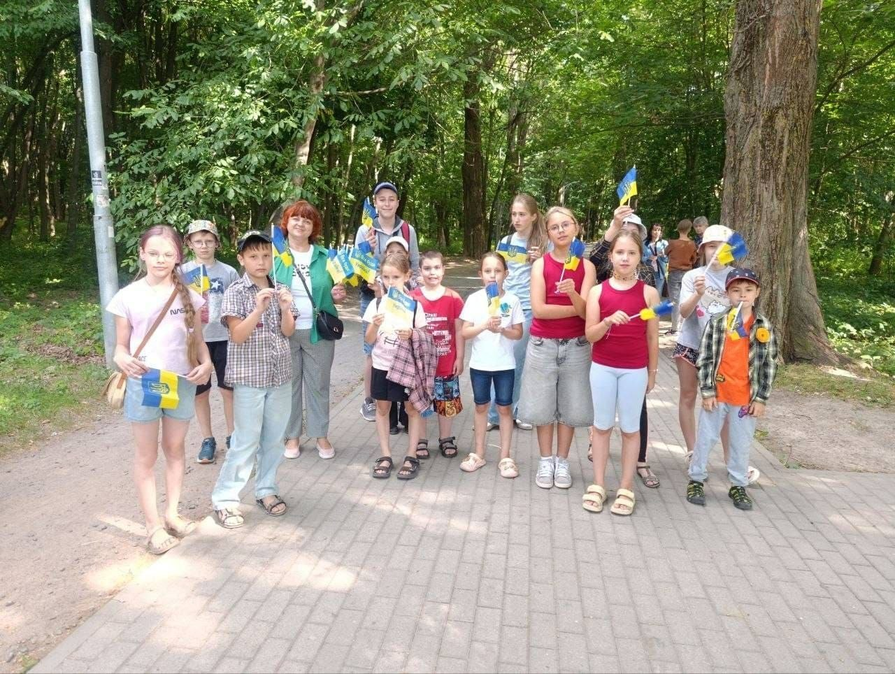 Група дітей та доросла жінка стоять на брукованій доріжці в парку, тримаючи в руках маленькі прапорці України та синьо-жовті кульки під час патріотичного заходу на свіжому повітрі.