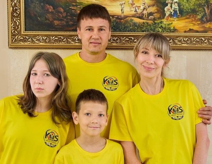 Портретне фото сім'ї з чотирьох осіб у однакових жовтих футболках із круглим логотипом «КМС» на тлі картини в золотистій рамі.