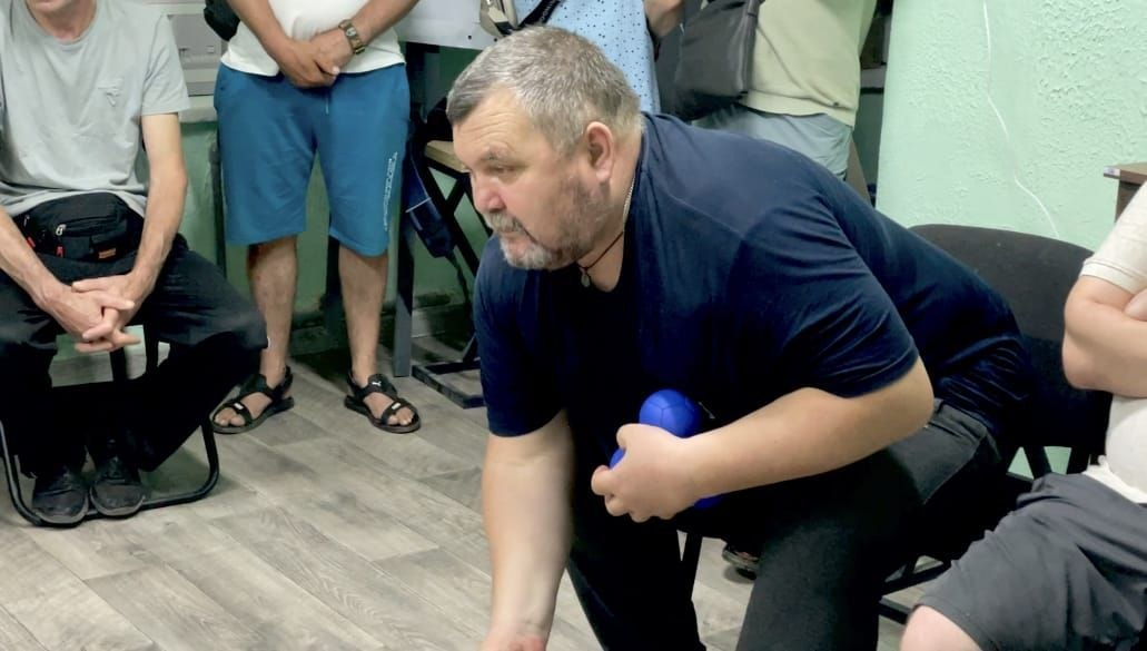 Чоловік у темній футболці нахиляється вперед, тримаючи в руці сині м'ячі для бочча, під час гри у приміщенні в оточенні інших людей.