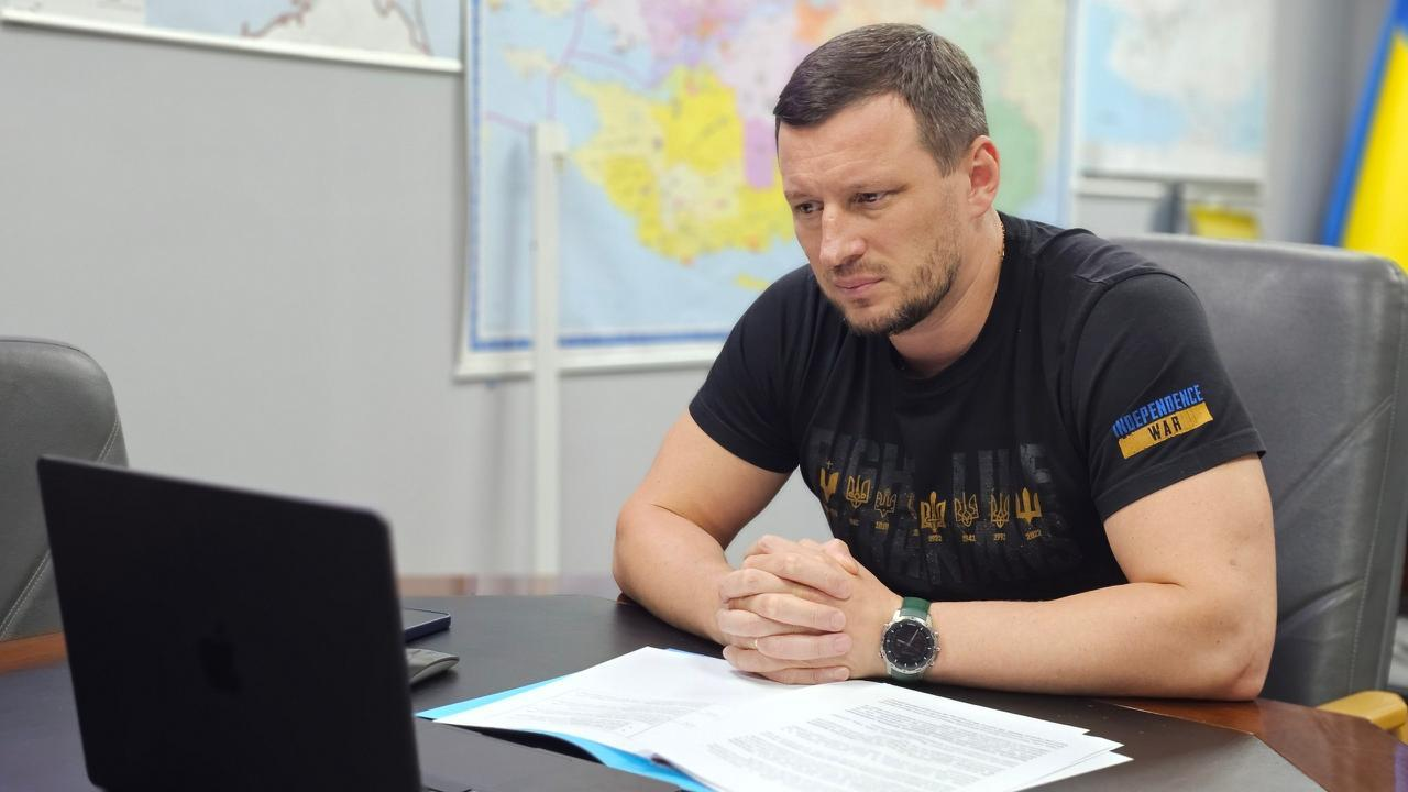 Чоловік у чорній футболці з українською символікою сидить за робочим столом перед ноутбуком та вивчає документи.