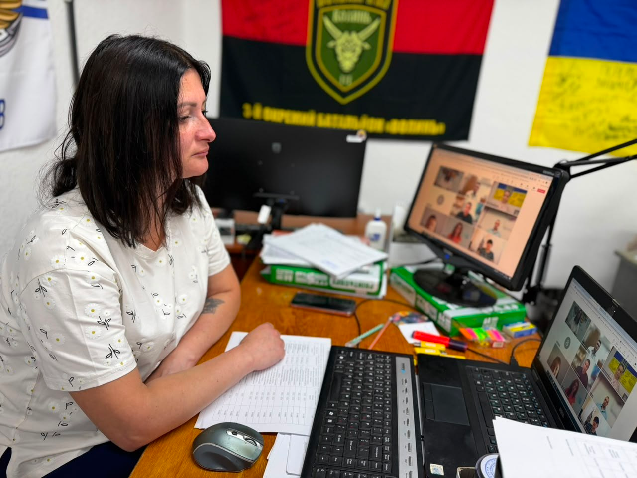 Жінка сидить за робочим столом біля комп'ютерних моніторів під час відеоконференції, на тлі червоно-чорного прапора з військовою символікою.
