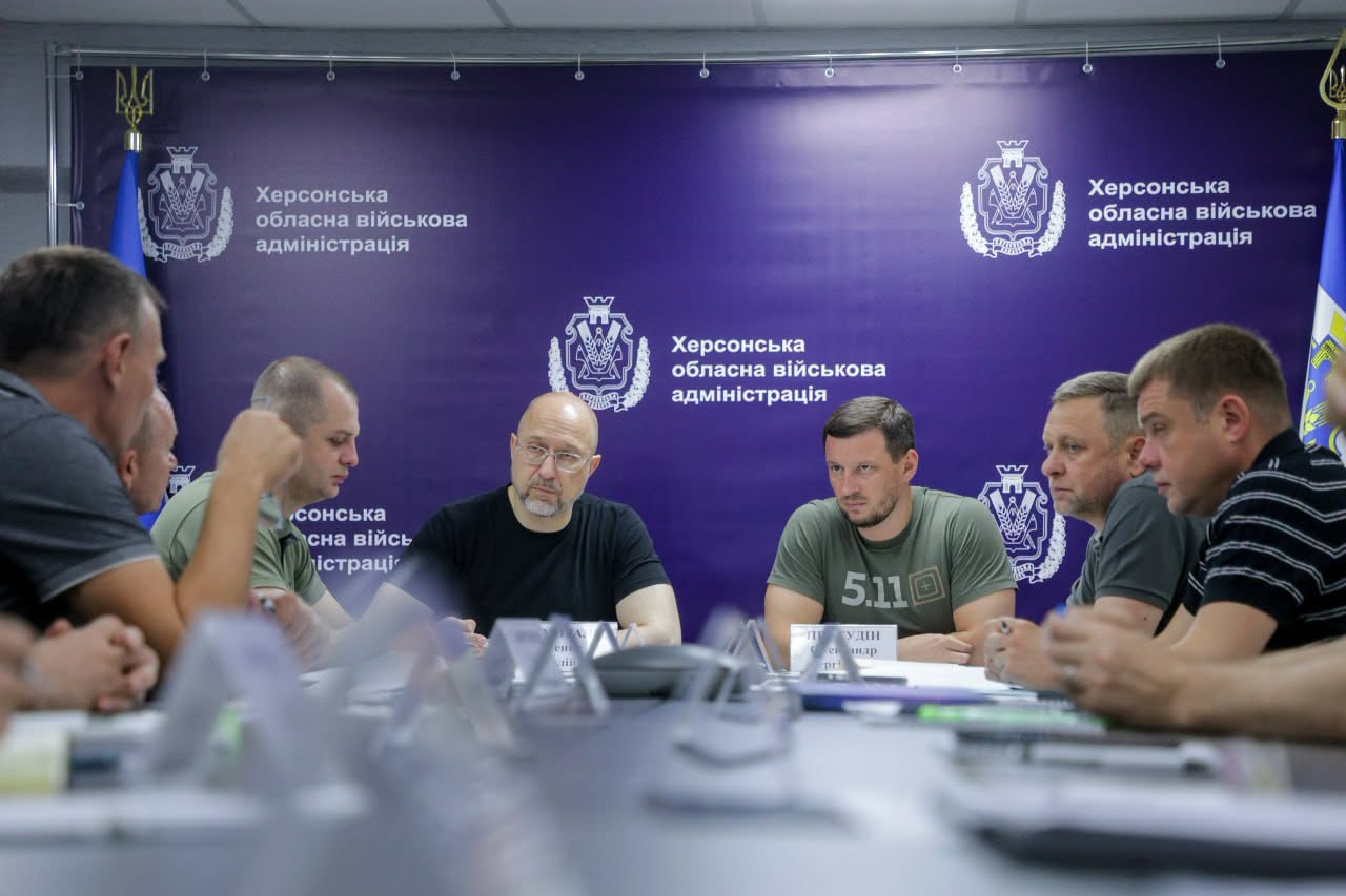 Група чоловіків сидить за столом переговорів під час офіційної наради на тлі великого фіолетового банера з гербами.