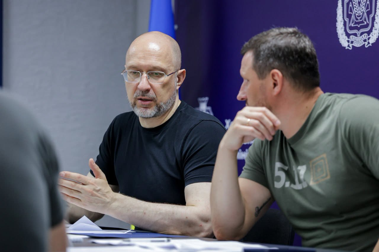 Чоловік в окулярах та чорній футболці жестикулює під час розмови з колегою за столом на офіційній зустрічі.