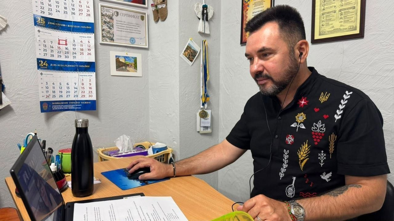 Чоловік у чорній сорочці з вишивкою сидить за робочим столом перед ноутбуком та тримає руку на комп'ютерній миші.