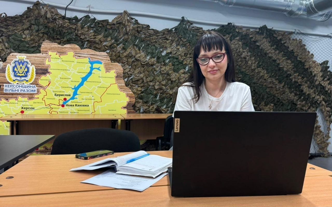 Жінка в окулярах сидить за робочим столом перед відкритим ноутбуком на тлі маскувальної сітки та стилізованої карти Херсонської області.