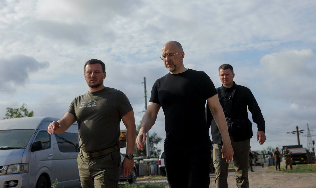 Трое чоловіків у повсякденному та тактичному одязі йдуть разом по відкритій місцевості на тлі хмарного неба та автомобілів.