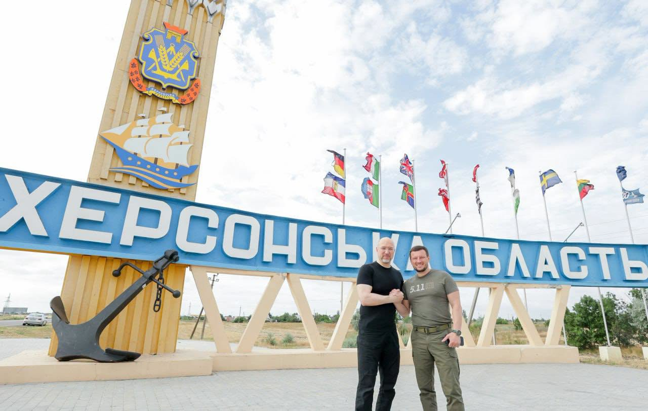 Два чоловіки стоять і тиснуть руки на тлі великої в'їзної стели з написом «Херсонська область», прикрашеної гербом, вітрильником та прапорами різних країн.