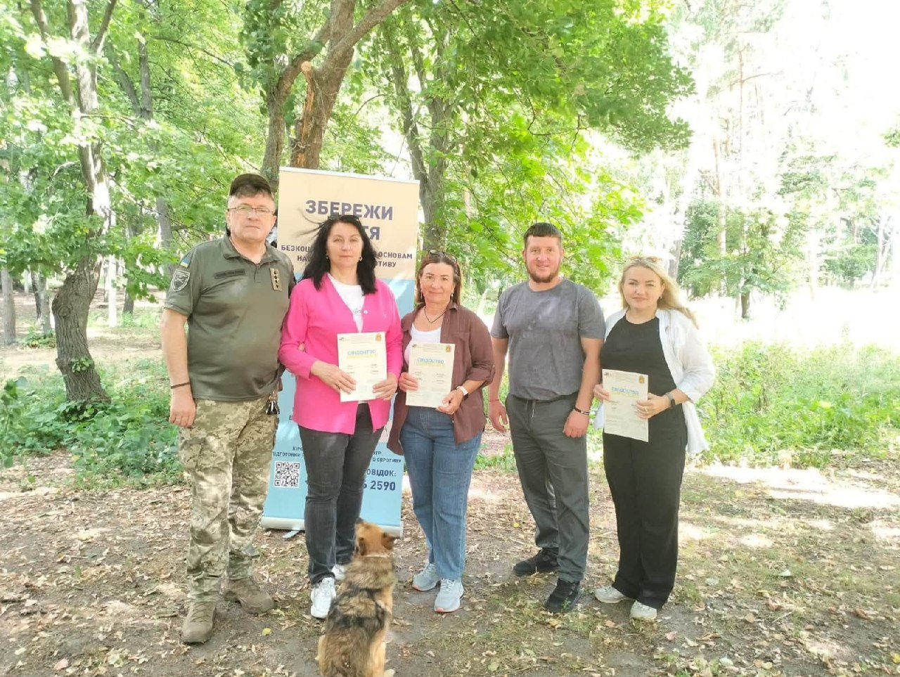 Група з трьох жінок та двох чоловіків стоїть у парку на тлі світлого інформаційного банера з написом «Збережи життя». Чоловік ліворуч одягнений у військову форму, інші люди — у повсякденний одяг, а троє учасниць тримають у руках паперові сертифікати. На передньому плані на землі спиною до камери сидить невеликий пухнастий собака.